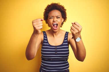 Beauitul Afro-Amerikan kadını izole edilmiş sarı arka plan üzerine yaz tişörtü giyer kızgın ve öfkeli yumruklar sallarken öfkeyle bağırır. Öfke ve agresif kavram.