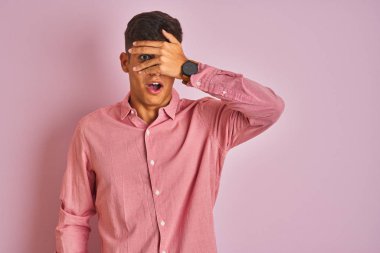 Young indian man wearing elegant shirt standing over isolated pink background peeking in shock covering face and eyes with hand, looking through fingers with embarrassed expression.