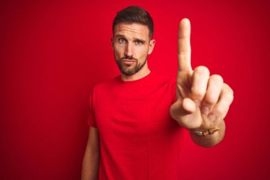 Kırmızı izole edilmiş arka plan üzerine gündelik tişört giyen genç yakışıklı adam parmağını kaldırmış ve kızgın bir ifadeyle işaret ediyor, hiçbir hareket göstermiyor.