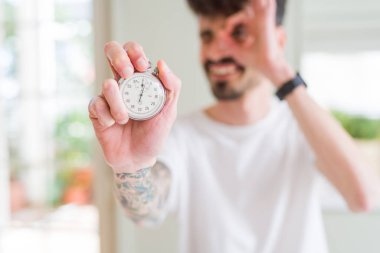 Genç adam elinde kronometreyle gülümsüyor. El pençe divan duruyor.