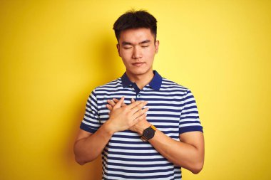Çizgili polo giyen Asyalı genç adam izole edilmiş sarı arka planda duruyor elleri göğsünde, gözleri kapalı ve yüzünde minnet dolu bir jest ile gülümsüyor. Sağlık kavramı.