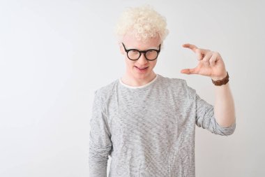 İzole edilmiş beyaz arka plan üzerinde çizgili tişört ve gözlük takan genç albino sarışın adam gülümsüyor ve el hareketleriyle küçük beden işareti yapıyor, parmakları görünüyor ve kameraya bakıyor. Ölçü kavramı.