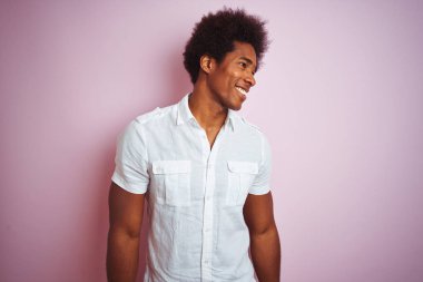 Afro saçlı, beyaz tişörtlü genç bir Amerikalı izole edilmiş pembe arka planda dikilip yüzünde gülümseme, doğal bir ifade ile yan yana bakıyor. Kendinden emin gülen.