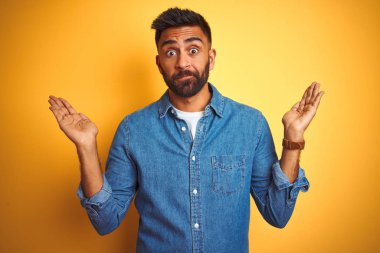 İzole edilmiş sarı arka plan üzerinde duran kot gömlekli genç Hintli adam. Kolları ve elleri havada, kafası karışmış ifade. Şüphe kavramı.