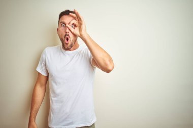Sürpriz yüz, göz parmaklarının arasından seyir ile ok jest yapıyor izole arka plan üzerinde rahat beyaz t-shirt giyen genç yakışıklı adam şok. İmansız ifade.