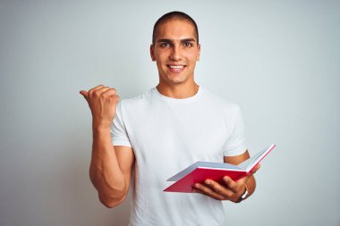 Genç yakışıklı adam beyaz izole edilmiş arka plan üzerinde kırmızı kitap okuyor ve gülümseyen bir gülümsemeyle baş parmağıyla işaret ediyor.
