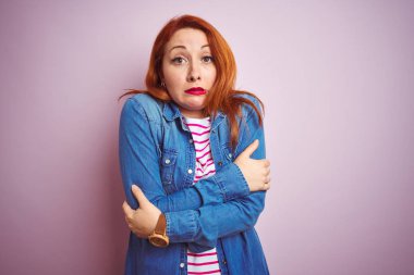 Güzel kızıl saçlı kadın kot gömlek ve çizgili tişört giyerek izole edilmiş pembe arka planda sallanıyor ve soğuktan donuyor. Yüzünde üzgün ve şok ifadesi var.