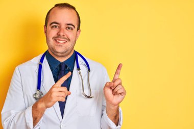 Ceket ve stetoskop giymiş genç bir doktor izole edilmiş sarı arka planda duruyor gülümsüyor ve kameraya bakıyor iki eliyle ve parmaklarıyla yan tarafa bakıyor..