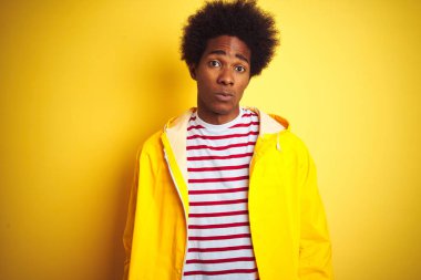 Afro saçlı Afrikalı bir adam izole edilmiş sarı arka planda duran şüpheli ve gergin bir yağmurluk giyiyordu. Sorunları yüzünden suratını asıyordu. Negatif kişi.