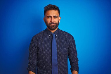 Zarif gömlek ve kravat giyen genç Hintli işadamı izole edilmiş mavi arka plan üzerinde duruyor şüpheci ve gergin, sorun yüzünden suratını asıyor. Negatif kişi.