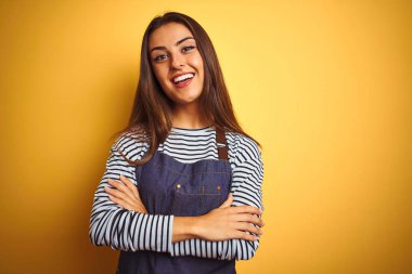 Önlük takan genç, güzel fırıncı kadın izole edilmiş sarı arka planda duruyor kollarını kavuşturup kameraya bakarken gülümsüyor. Pozitif kişi.