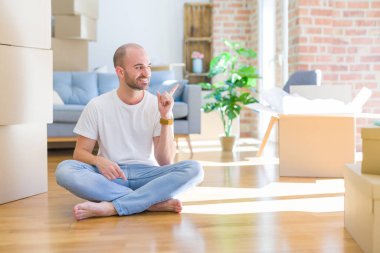 Genç kel adam, karton kutuların etrafında oturuyor. Yüzünde kocaman bir gülümsemeyle yeni bir eve taşınıyor. Elleri ve parmaklarıyla kameraya bakıyor..