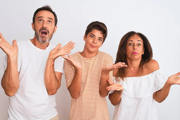 Family of three, mother, father and son standing over white isolated background clueless and confused expression with arms and hands raised. Doubt concept.