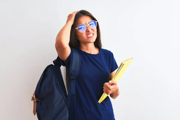 Young student woman wearing backpack glasses holding book over isolated white background stressed with hand on head, shocked with shame and surprise face, angry and frustrated. Fear and upset for mistake.