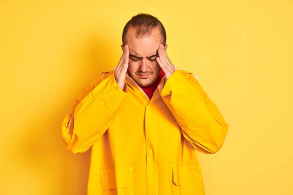 Young man wearing rain coat standing over isolated yellow background with hand on head for pain in head because stress. Suffering migraine.
