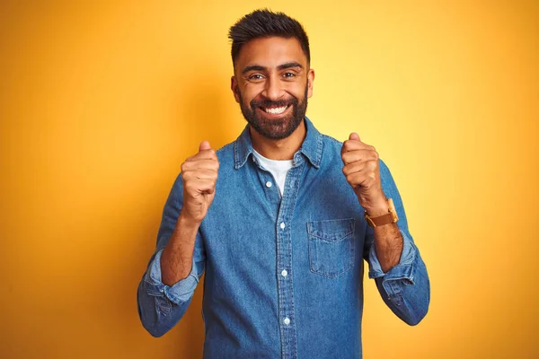 独立した黄色の背景の上に立つデニムシャツを着た若いインド人男性が腕を上げて成功に興奮し 目は勝利笑顔を祝う閉じました 勝者のコンセプト — ストック写真
