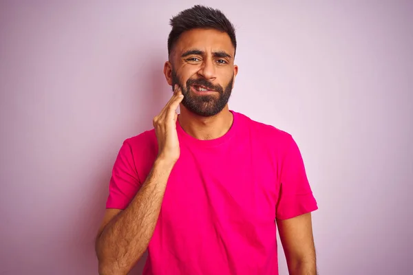 Young indian man wearing t-shirt standing over isolated pink background touching mouth with hand with painful expression because of toothache or dental illness on teeth. Dentist concept.