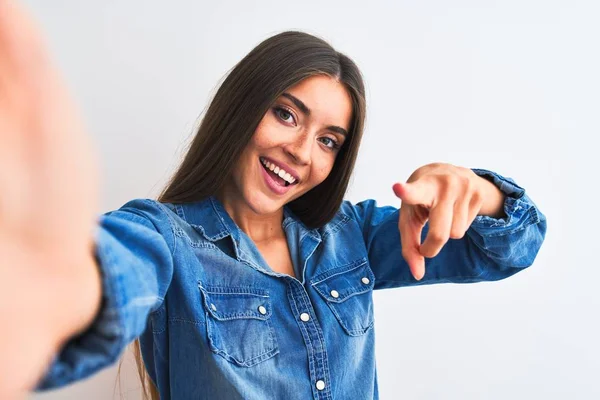 Beautiful woman wearing denim shirt make selfie by camera over isolated white background pointing to you and the camera with fingers, smiling positive and cheerful