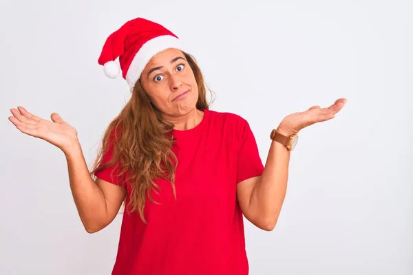 孤立した背景の上にクリスマスの帽子を身に着けている中年の成熟した女性は腕と手で上げられ混乱した表現 疑いの概念 — ストック写真