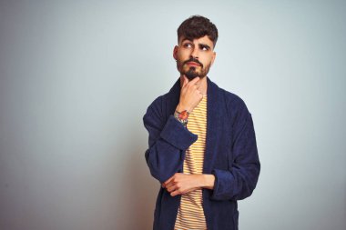 Dövmeli, bornozlu genç bir adam, soyutlanmış beyaz arka planda duruyor, elleri çenesinde soru sorarken, düşünceli bir ifade takınıyor. Düşünceli bir yüzle gülümsüyor. Şüphe kavramı.