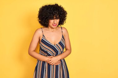 Kıvırcık saçlı, çizgili renkli elbiseler giyen Arap bir kadın. İzole edilmiş sarı arka plan, mide bulantısı, acı verici bir hastalık yüzünden el üstünde. Ache kavramı.