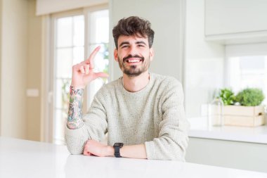 Genç bir adam beyaz bir masada oturmuş gülümsüyor ve kendine güvenen hareketler yapıyor parmaklarıyla işaret yapıyor ve kameraya bakıyor. Ölçü kavramı.