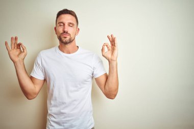 İzole edilmiş arka planda günlük beyaz tişört giyen genç yakışıklı adam rahatlamış ve parmaklarıyla meditasyon hareketi yaparken gözleri kapalı gülümsüyor. Yoga konsepti.