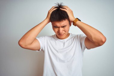 Asyalı genç bir Çinli tişört giyiyor. İzole edilmiş beyaz arka plan üzerinde duruyor. Ağrı ve migren yüzünden baş ağrısı ve stresten muzdarip. Ellerini başının üstüne koy..