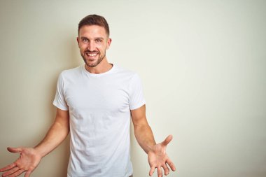 İzole edilmiş arka planda günlük beyaz tişört giyen genç yakışıklı adam güler yüzlü, kolları açık, dostça karşılama, olumlu ve kendinden emin selamlar.