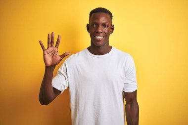 Beyaz t-shirt giyen genç Afro-Amerikalı adam izole edilmiş sarı arka plan üzerinde duruyor ve beş numarayı gösterirken kendinden emin ve mutlu gülümsüyor..