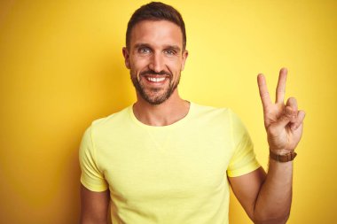 Sarı izole edilmiş arka plan üzerine gündelik sarı tişört giyen genç yakışıklı adam kameraya gülümseyip zafer işareti yapıyor. İki numara..