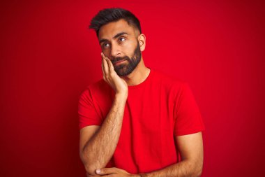 Genç, yakışıklı Hintli bir adam, izole edilmiş kırmızı arka planda tişört giyiyor. Kollarını kavuşturup depresyondan sıkılmış ve yorgun görünüyor..