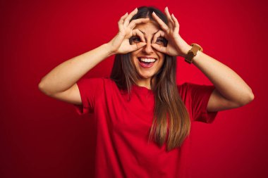 İzole edilmiş kırmızı arka plan üzerinde duran tişört giyen genç ve güzel bir kadın dürbün gibi güzel hareketler yapıyor. Dürbün gibi dilini dışarı çıkarıyor, gözler parmaklarından bakıyor. Çılgın ifade.