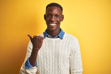 İzole edilmiş sarı arka plan üzerinde kot gömlek ve beyaz süveter giyen, gülümseyen ve baş parmağıyla yüzünü gösteren Afro-Amerikan bir adam..