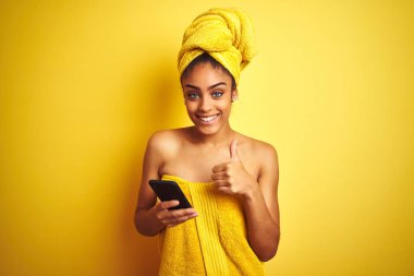 Duş üstüne havlu giyen Afro kadın izole edilmiş sarı arka plan üzerinde smatrphone kullanıyor. Gülümsüyor ve gülümsüyor.
