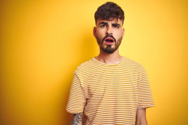 İzole edilmiş sarı arka plan üzerinde duran çizgili tişörtlü genç adam. Şok suratlı, şüpheci ve iğneleyici bakışlı, ağzı açık şaşırmış.