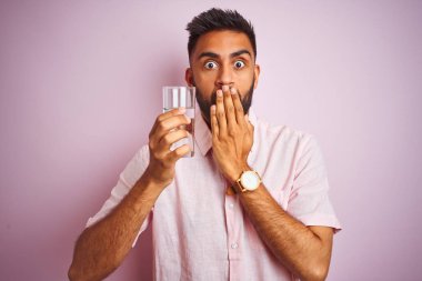 Genç, yakışıklı Hintli bir adam, izole edilmiş pembe arka planda bir bardak su içiyor. Ağzı kapalı. Yanlışlıkla utançla sarsılan ağzı, korkunun ifadesi, sessizlikten korkan gizli konsept.