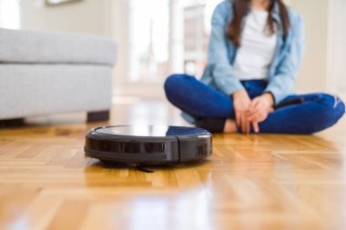 Yerleri temizlemek için otomatik süpürge kullanan genç bir kadın makine ev işi robotu kontrol ediyor.