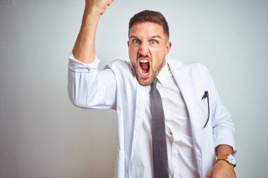 Genç, yakışıklı doktor, izole edilmiş arka planda beyaz bir palto giyiyor kızgın ve kızgın. Öfkeyle bağırırken yumruğunu kaldırıyor. Öfke ve agresif kavram.