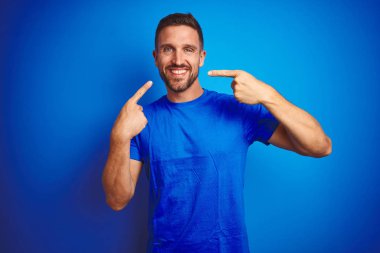 Mavi izole edilmiş arka planda günlük tişört giyen genç yakışıklı adam gülümsüyor ve parmaklarıyla ve ağzıyla işaret ediyor. Diş sağlığı konsepti.