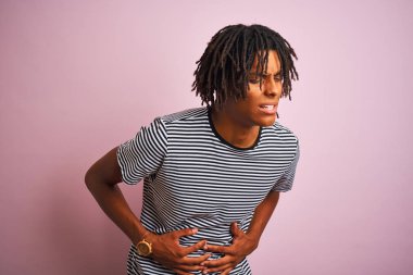 Rasta desenli tişörtlü Afro bir adam midesinde hazımsızlık, acı veren bir hastalık yüzünden izole edilmiş pembe arka planda dikiliyor. Ache kavramı.