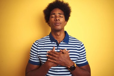 Afro saçlı, donanma çizgili polo giyen Amerikalı adam izole edilmiş sarı arka planda duruyor elleri göğsünde, gözleri kapalı ve yüzünde minnet dolu bir jest ile gülümsüyor. Sağlık kavramı.