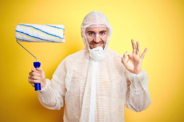 Boyama ekipmanı giyen ve sarı arka planda boya silindiri tutan genç adam parmaklarıyla tamam işareti yapıyor, mükemmel bir sembol.