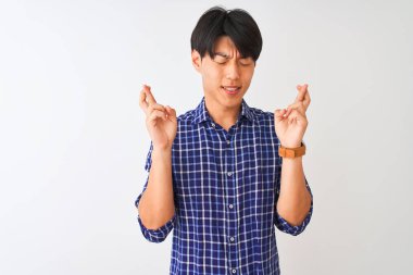 İzole edilmiş beyaz arka plan üzerinde dikilen sıradan mavi gömlekli genç Çinli adam umut ve gözler kapalı bir şekilde gülümseyerek işaret ediyor. Şans ve batıl inanç..