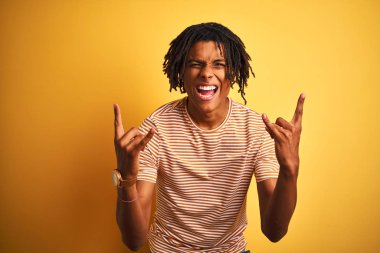 Rastalı tişörtlü Afro adam izole edilmiş sarı arka plan üzerinde duruyor çılgın bir ifadeyle elleri havada rock sembolü yapıyor diye bağırıyor. Müzik yıldızı. Ağır müzik konsepti.