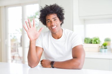 Genç Afro-Amerikalı adam, günlük beyaz tişörtler giyip evde oturuyor ve parmaklarını beş numarayı göstererek gülümsüyor ve gülümsüyor..