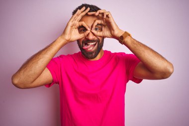 İzole edilmiş pembe arka plan üzerinde duran genç Hintli adam dürbünün dili dışarı çıkarması, gözlerin parmaklarından içeri bakması gibi güzel hareketler yapıyor. Çılgın ifade.