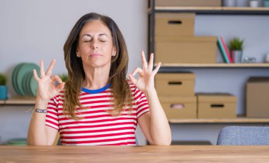 Orta yaş yaşlı kadın evde masada oturan rahat ve gözleri parmakları ile meditasyon jest yapıyor kapalı gülümseyerek. Yoga konsepti.