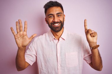 İzole edilmiş pembe arka plan üzerinde duran genç Hintli adam kendini beğenmiş ve mutlu bir şekilde gülümserken altı numaralı parmaklarıyla işaret ediyor..