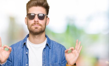 Genç yakışıklı zarif adam giyen denim ceket rahatla ve gülümseyen gözlerle kapalı parmak jestle meditasyon yapıyor. Yoga kavramı.